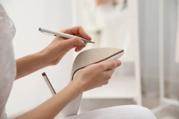 Photo of Young bride writing on her shoe indoors, closeup. Wedding superstition