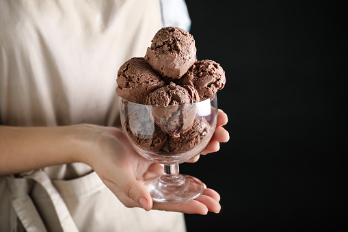 Woman holding glass bowl full of chocolate ice cream on black background, closeup. Space for text