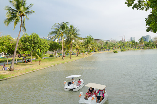 View over lake in park Chatuchak in Bangkok. People are relaxing in park and on pedo boats on lake. In background is some skyline and BTS skytrain station MoChit