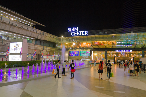 People are at night between Siam Center and SIam Paragon malls and BTS skytrain at night in Bangkok at Sukhumvit Road. People are walking between malls. Some are standing at illuminated fountain. In ;eft background is BTS skytrain