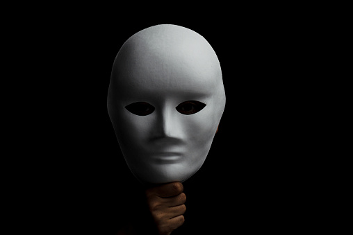 Person is hiding face behind a white mask on black background.