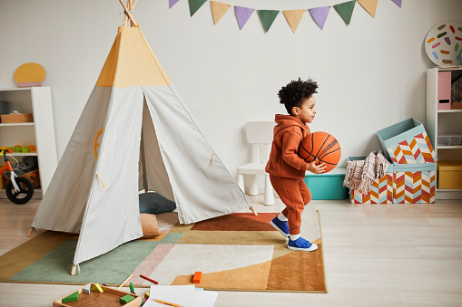 Full length portrait of cute African American boy holding basketball ball while playing at home in cozy kids room and wearing minimal trendy outfit