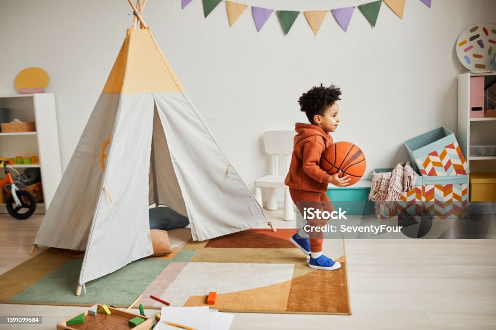 Little Boy in Cute Playroom Full length portrait of cute African American boy holding basketball ball while playing at home in cozy kids room and wearing minimal trendy outfit Basketball - Sport Stock Photo