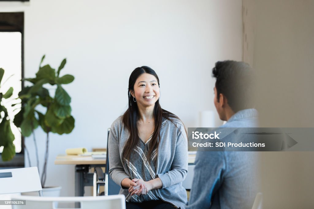 Mid adult female business owner smiles at job applicant The mid adult female business owner clasps her hands and smiles as she listens to the unrecognizable young adult male job applicant. Meeting Stock Photo