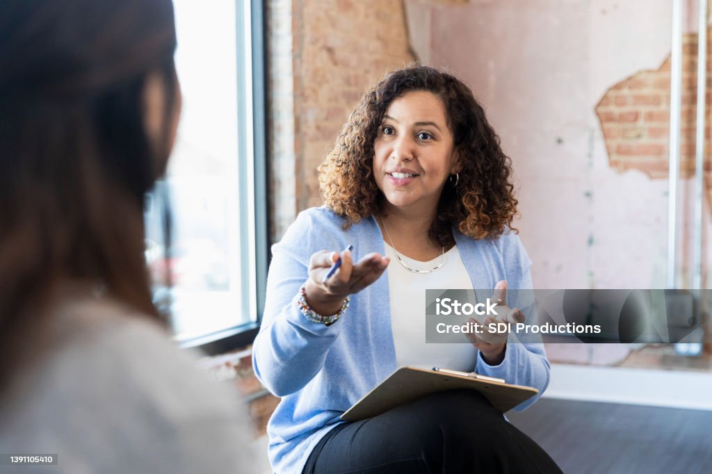 Meeting one-on-one, female therapist gestures toward unrecognizable woman Meeting with her client one-on-one, the mid adult counselor gestures toward her unrecognizable female client. Human Resources Stock Photo