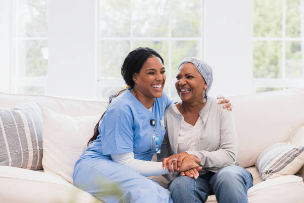 Home health nurse and female patient embrace and laugh together During her visit, the mid adult home health nurse and the senior adult woman with cancer embrace and laugh together. home caregiver stock pictures, royalty-free photos & images
