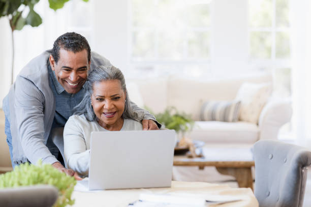 el esposo mira por encima del hombro de la esposa las fotos de los nietos en una computadora portátil - senior couple fotos fotografías e imágenes de stock