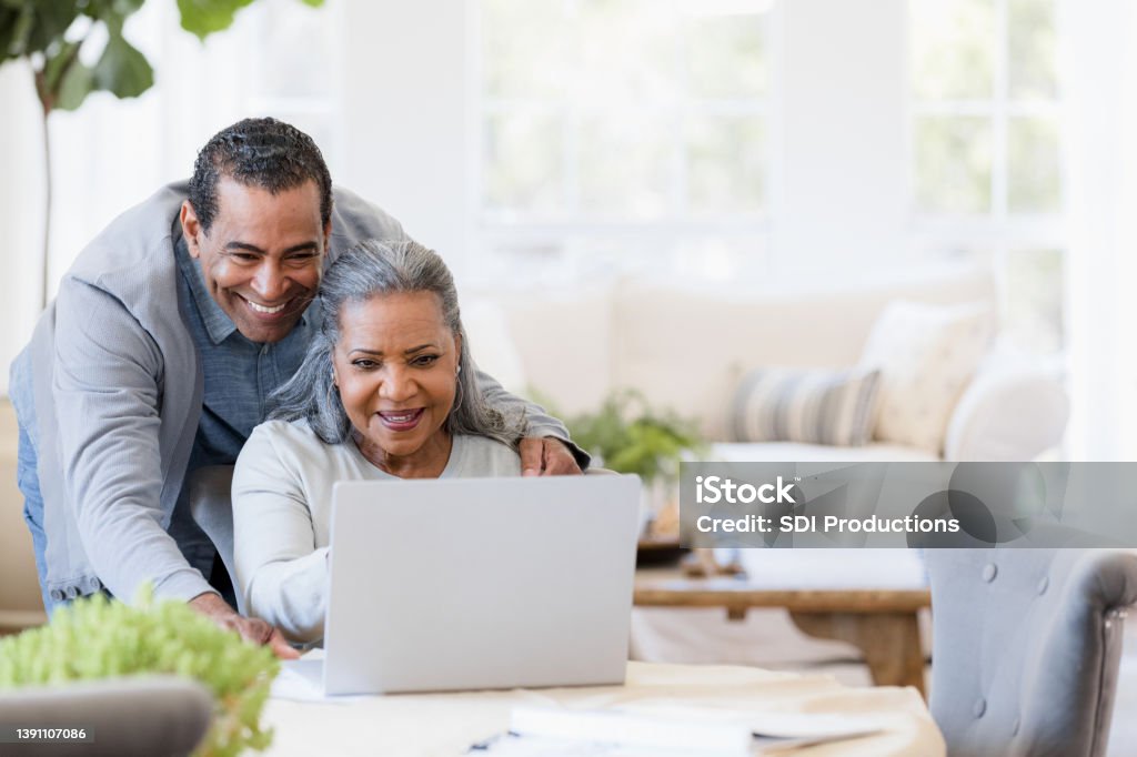 Husband looks over wife's shoulder at grandchildren's photos on laptop The senior adult husband smiles broadly as he looks over his wife's shoulder at the grandchildren's photos displayed on the laptop screen. Senior Couple Stock Photo