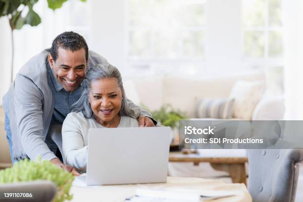 Husband Looks Over Wifes Shoulder At Grandchildrens Photos On Laptop Stock Photo - Download Image Now