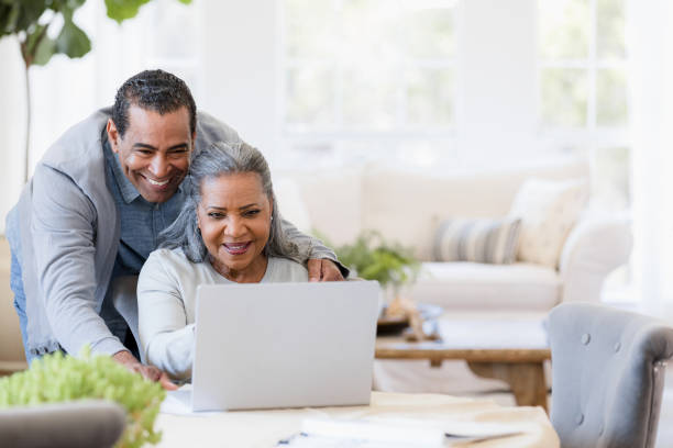 husband looks over wife's shoulder at grandchildren's photos on laptop - couple smiling happiness people imagens e fotografias de stock