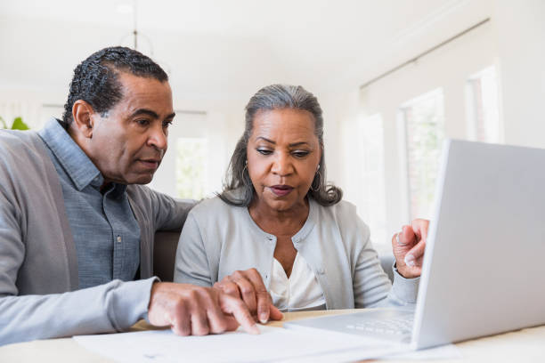 пожилая пара сравнивает бумажную работу с онлайн-записями - home finances couple computer african ethnicity стоковые фото и изображения