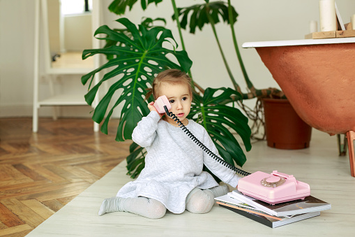 A cute little girl is sitting on the floor next to the bathtub and the monstera plant and talking on an old vintage pink push-button telephone apparatus. A child plays with a handset