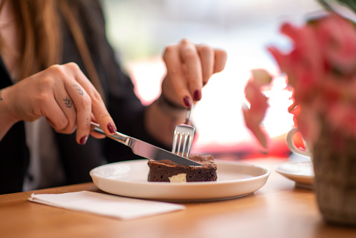 Young woman eating chocolate cake with a cup of coffee in a coffee shop. She is cutting a slice of cake with a knife.