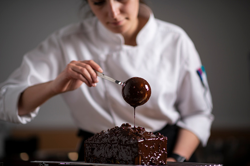 Female baker pouring chocolate sauce on cake with ladle.