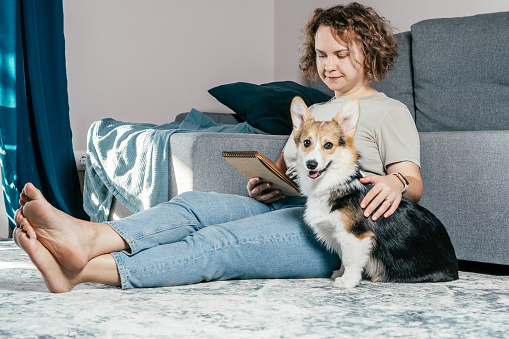 Curly haired glad, cheerful barefoot woman with dog corgi sitting, relaxing and reading electronic book on carpet in comfortable living room near sofa. Calm and cozy atmosphere. Weekend vacation