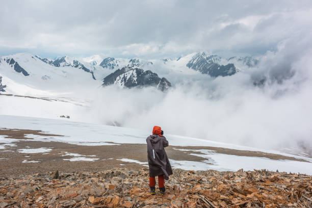 istock Hiker and high snowy mountains in dense low clouds. Minimal scenery with man on stone hill against beautiful mountain peaks in thick clouds. Simple minimalism with man and majestic mountains nature. 1391129589