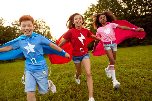 Company of multiracial delighted kids in superhero capes running on lawn in summer and enjoying freedom