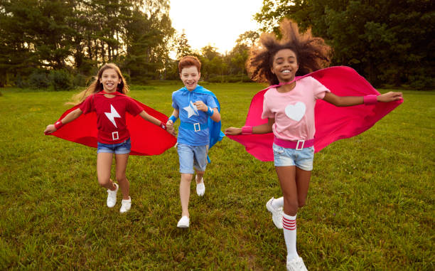 Cheerful little superheros running in park Happy diverse kids in superhero costumes smiling and running on grassy lawn while playing together in park at sunset cape garment stock pictures, royalty-free photos & images