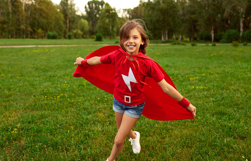 Delighted girl in red superhero cape running along meadow in park while looking at camera and having fun in summer