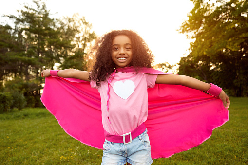 Positive African American preteen girl wearing pink superhero cape standing on lawn in park and looking at camera while having fun