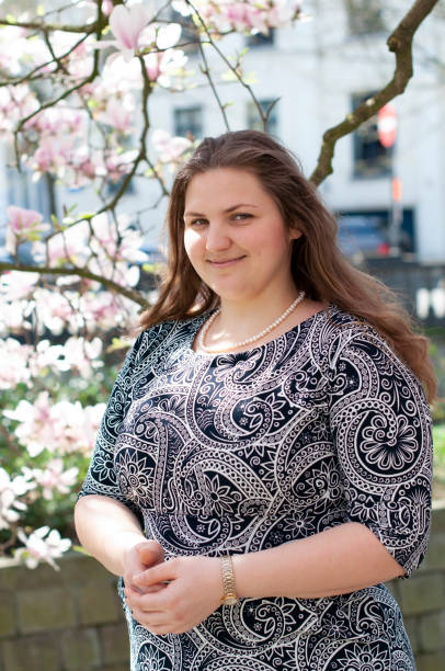 Portrait of a beautiful plump caucasian confident woman A plump, confident woman stands under a blooming magnolia and smiles.    young millennial woman with brown curly hair smiling and waiting for a friend for a date. A full girl enjoys flowering and spring. High quality photo magnolia white flower large stock pictures, royalty-free photos & images