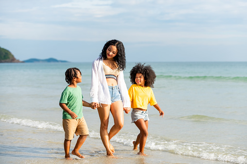 Happy African family mother holding hands with little daughter and son walking together on the beach at summer sunset. Mom with child boy and girl enjoy and fun outdoor lifestyle vacation at the sea