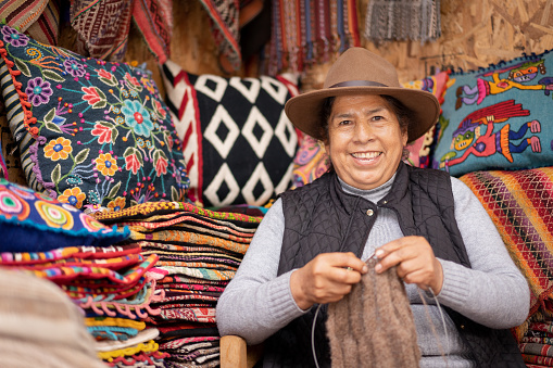 Peruvian woman weaving baby alpaca wool in a handicraft store with ruanas and colorful cushions in the background.