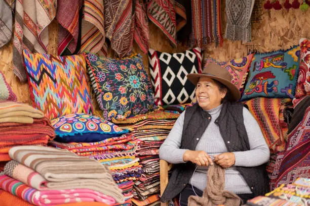 Peruvian woman weaving baby alpaca wool in a handicraft store with ruanas and colorful cushions in the background.