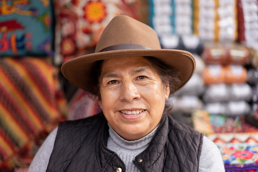 Portrait of a Peruvian woman wearing a typical hat in an alpaca wool handicraft shop, with ruanas and cushions in the background.