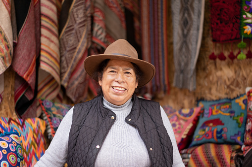 Portrait of a Peruvian woman wearing a typical hat in an alpaca wool handicraft shop, with ruanas and cushions in the background.