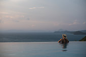 Rear view of a woman with hat relaxing in infinity swimming pool and looking at a beautiful sunset and the sea view
