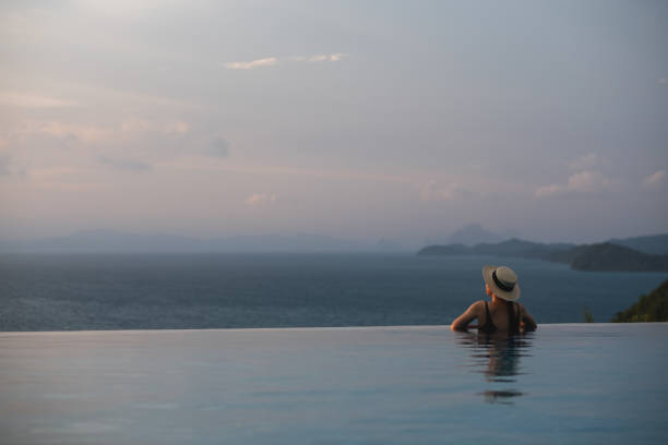 Rear view of a woman with hat relaxing in infinity swimming pool and looking at a beautiful sunset and the sea view Rear view of a woman with hat relaxing in infinity swimming pool and looking at a beautiful sunset and the sea view infinity pool stock pictures, royalty-free photos & images