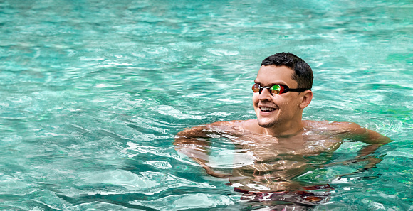 Swimmer. Side view of smiling athletic man in swimming goggles in pool. Panorama banner. Healthy lifestyle. Young athlete on the background of turquoise water. Sports and fitness cardio exercises.
