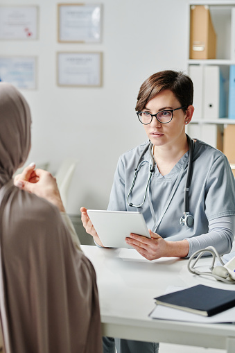 Young serious clinician with tablet consulting Muslim female patient in hijab sitting in front of her and giving her medical advice