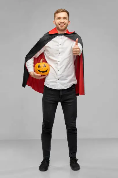 holiday, theme party and people concept - happy smiling man in halloween costume of vampire with dracula cape showing thumbs up and holding jack-o-lantern pumpkin over grey background
