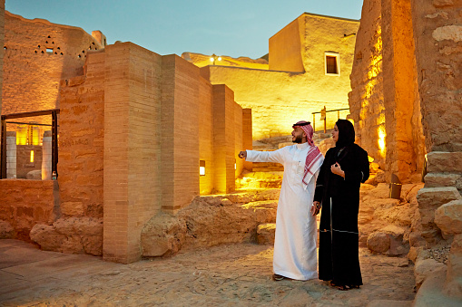 Full length view of man and woman in their 30s wearing traditional Saudi attire and admiring features of illuminated open air museum. Property release attached.