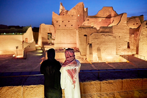 Rear view of man and woman in traditional attire admiring illuminated Salwa Palace, original home of Saudi royal family and part of At-Turaif ruins. Property release attached.