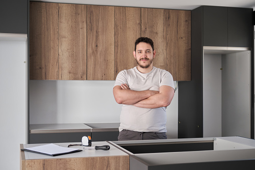 Young hispanic man smile and cross his arms after installing furniture on modern new kitchen.