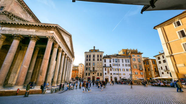 A suggestive glimpse of the square of the Roman Pantheon in the historic heart of Rome Rome, Italy, February 03 -- A suggestive glimpse of Piazza della Rotonda and the Roman Pantheon, in the historic heart of the Eternal City, with some tourists lining up to visit the famous temple and the typical outdoor tables of Italian restaurants. Built in 27 BC by the Consul Marco Vispanio Agrippa for the emperor Augustus and dedicated to the Roman gods, the majestic Pantheon is one of the best preserved Roman structures in the Eternal City and in the world. The fountain in the center of the square (right), with a real Egyptian obelisk, was built by the sculptor Leonardo Sormani in 1575 on a project by the architect Giacomo della Porta. In 1980 the historic center of Rome was declared a World Heritage Site by Unesco. Super wide angle image in 16:9 and high definition format. ancient rome photos stock pictures, royalty-free photos & images