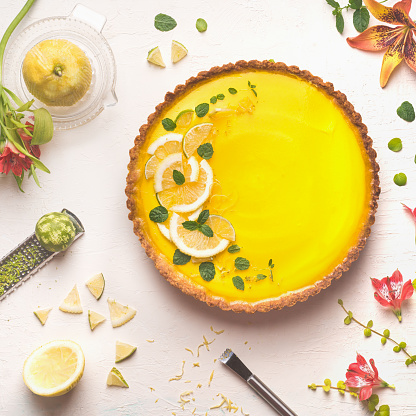 Yellow lemon tart topped with fresh lemon and lime slices on white table background with citrus ingredients and flowers, top view. Traditional french cuisine