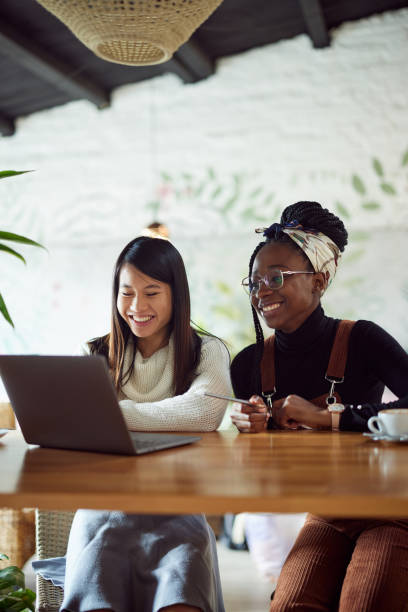 Due ragazze universitarie multiculturali in piedi nella caffetteria e che usano un laptop per la ricerca. E-learning e disponibilità dei dati. - foto stock