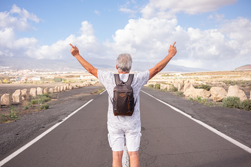 Back view of senior caucasian man with backpack on shoulders walking in empty street outdoors gesturing peace or victory with open arms. White-haired old man enjoying freedom and nature. Copy space