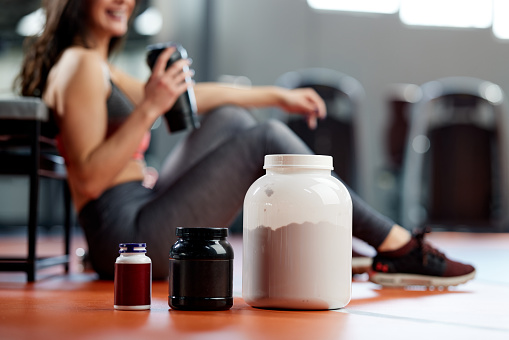 Selective focus on protein powder and multivitamins jars on the gym floor with a sporty woman in background.