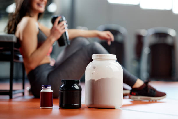 Selective focus on protein powder and multivitamins jars on the gym floor with a sporty woman in background. stock photo