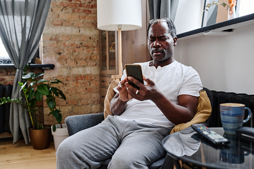 Contemporary serious old man with smartphone looking at its screen while texting or reading message during home rest