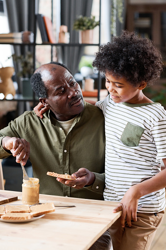 Cute multi-ethnic schoolboy standing by his grandfather making him sandwich with peanut butter for breakfast by wooden table