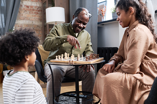 Happy retired black man pointing at pawn on chess board while explaining his grandchildren rules of leisure game