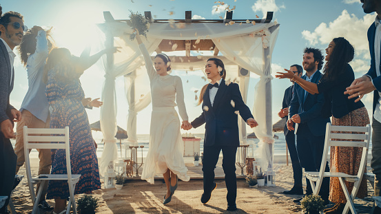 Attractive Female Queer Couple Walking Up the Aisle at Outdoors Wedding Ceremony Near Ocean. Two Lesbian Women in Love Share Happiness with Diverse Multiethnic Friends. Cute LGBTQ Relationship Goals.