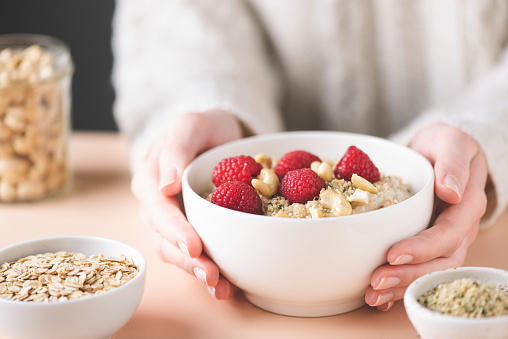 Oatmeal bowl with raspberry, cashew nuts and hemp seeds in female's hands. Clean eating, dieting concept