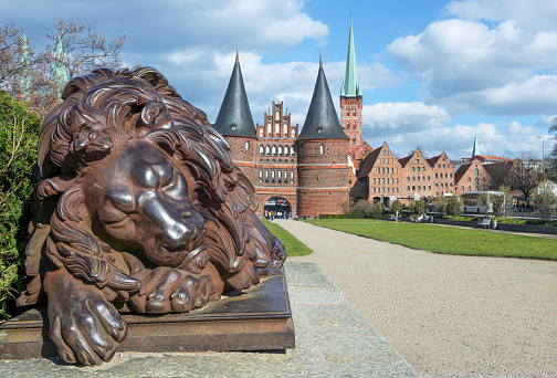 Sleeping lion sculpture from iron in front of the Holsten gate or Holstentor, the landmark of the historic hanseatic city of Lubeck in Germany, blue sky with clouds, copy space